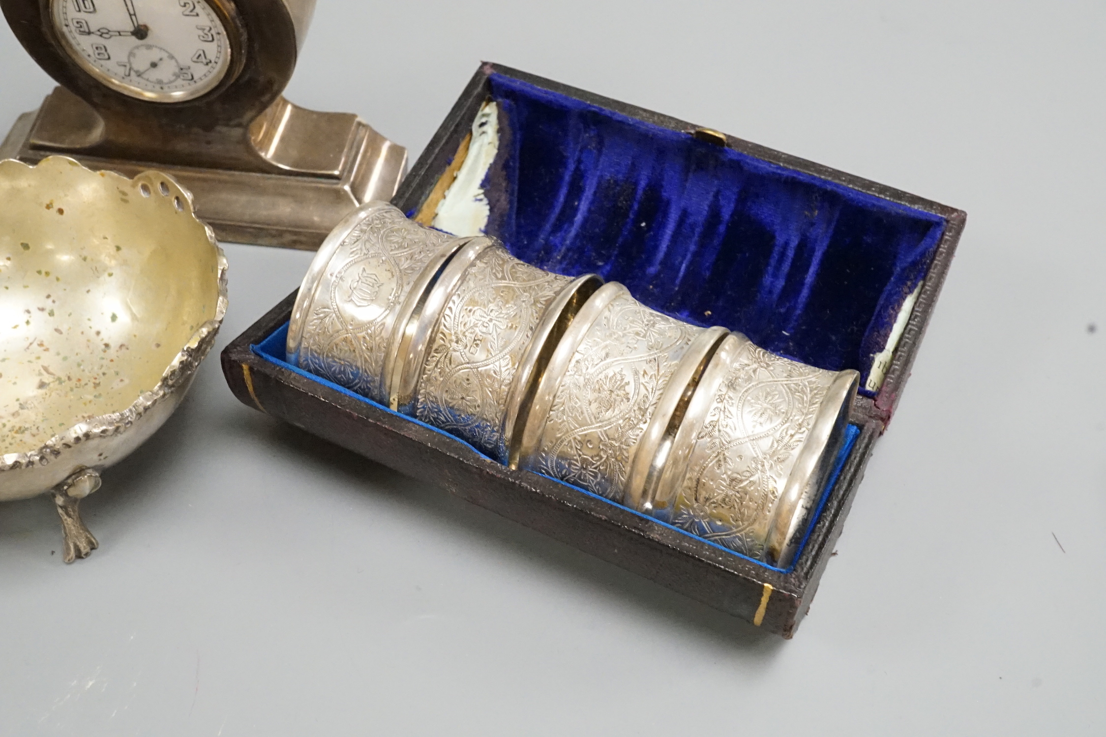 A George V silver mounted small mantel clock, height 93mm, a pair of silver pepperettes and eight other items including a silver mounted cigarette box, a cased set of four chased silver napkin rings and two plated hip fl